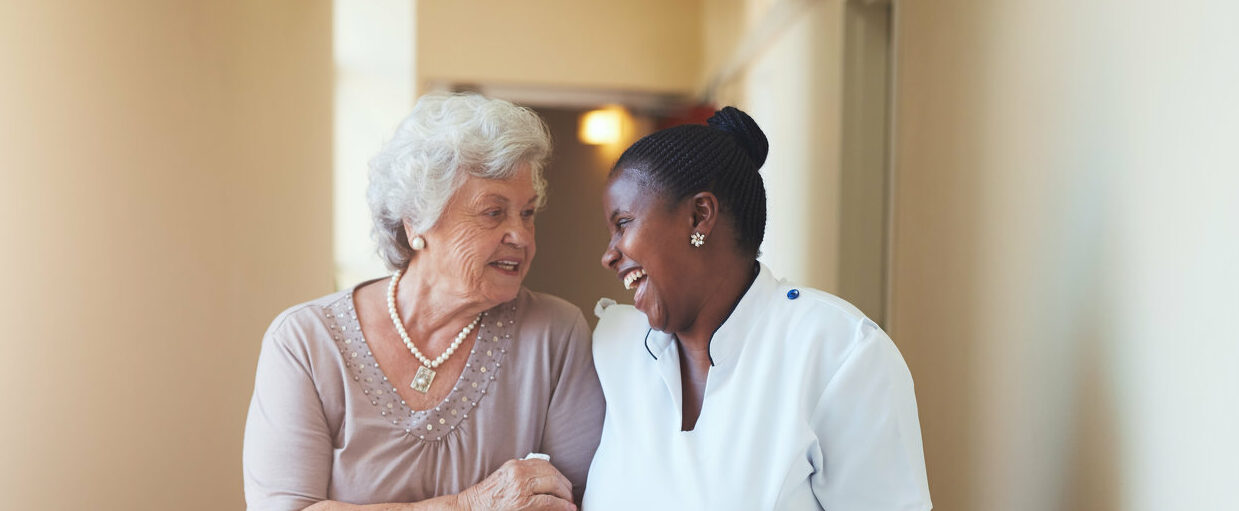 Portrait of happy female caregiver and senior woman walking together at home. Professional caregiver taking care of elderly woman. 2022/03/AdobeStock_112047591-e1647986317495.jpeg 