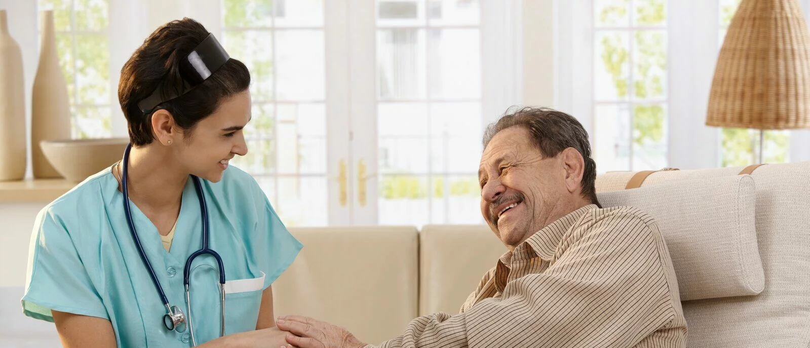 Happy nurse holding hands of elderly patient sitting side by side at home, laughing. 2022/03/AdobeStock_24969857-e1647892175946.jpeg 